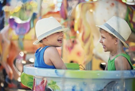 Two little brothers sitting in the same cup on carousel. The boys are aged 5 and wearing fedoras. Boys are having fun and laughing.